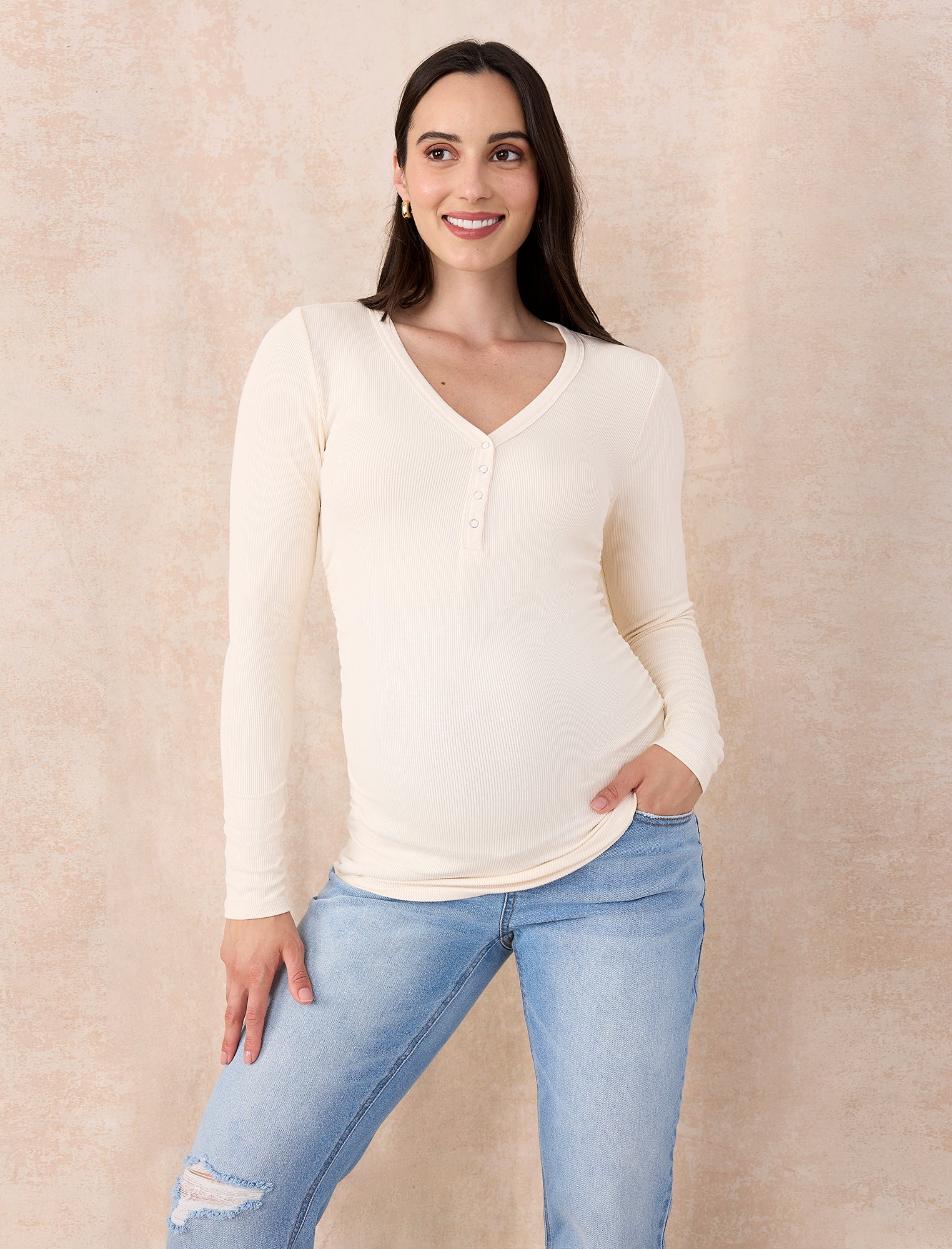 Pregnant woman wearing a cream long-sleeve top and light blue jeans against a beige background