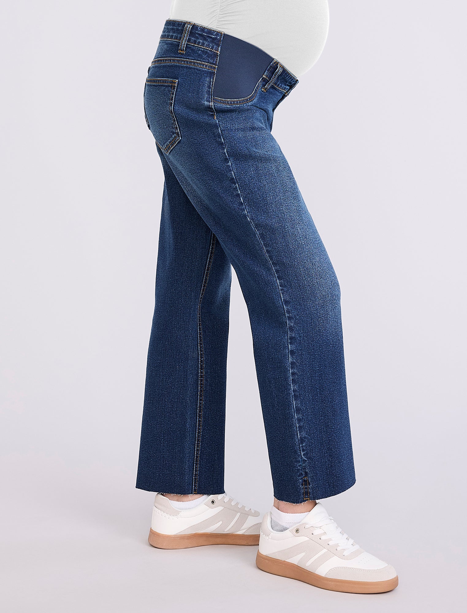 Person wearing maternity blue jeans and white sneakers on a light gray background