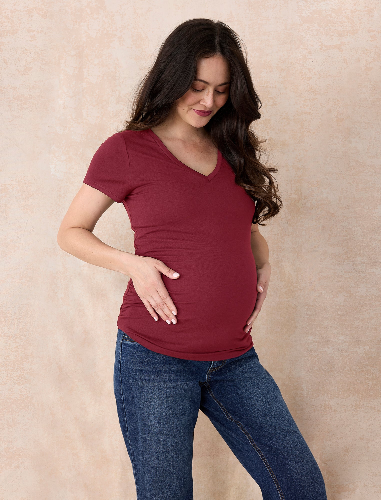Pregnant woman wearing a maroon t-shirt and jeans against a plain background