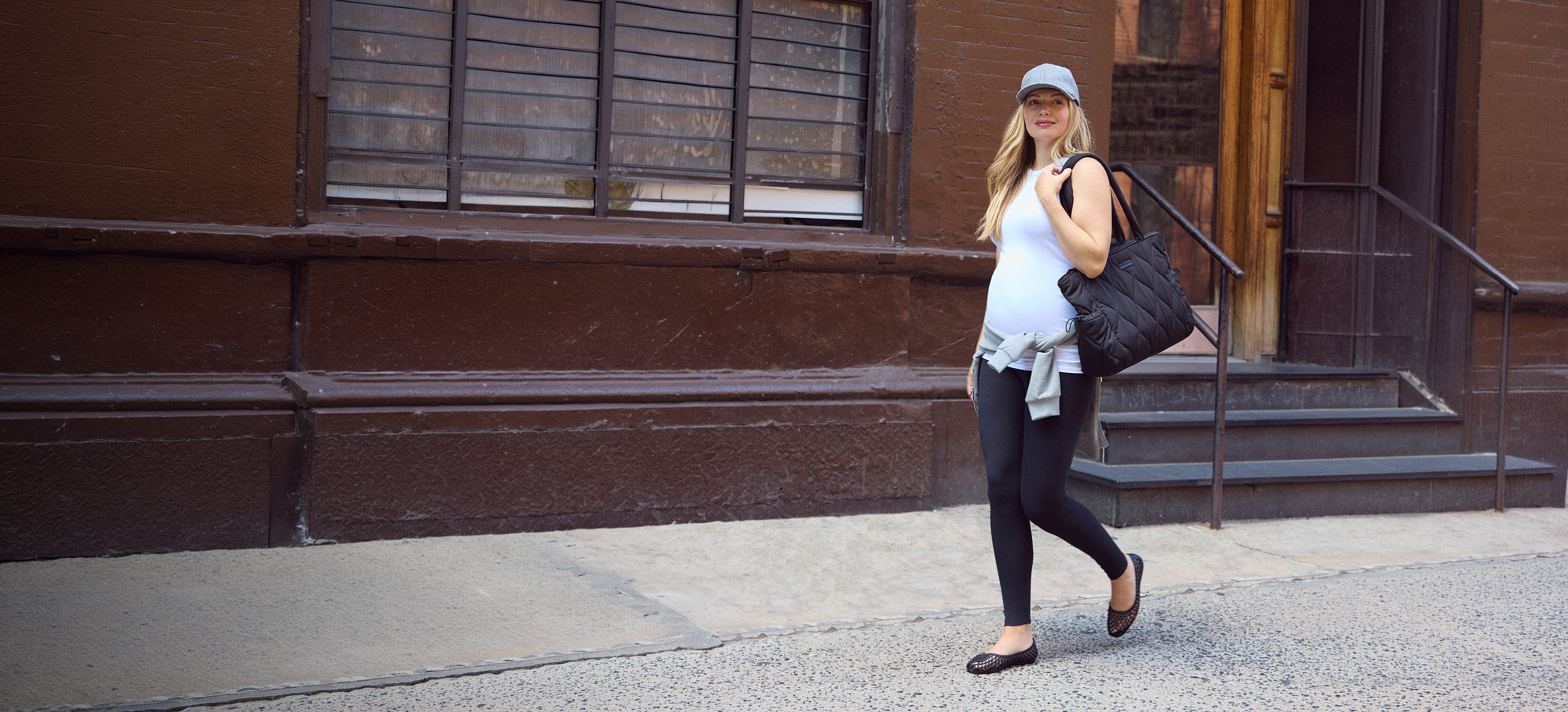 A woman walking on a city sidewalk in front of a brown brick building, wearing a white sleeveless top, black leggings, dark flats, and carrying a black shoulder bag.
