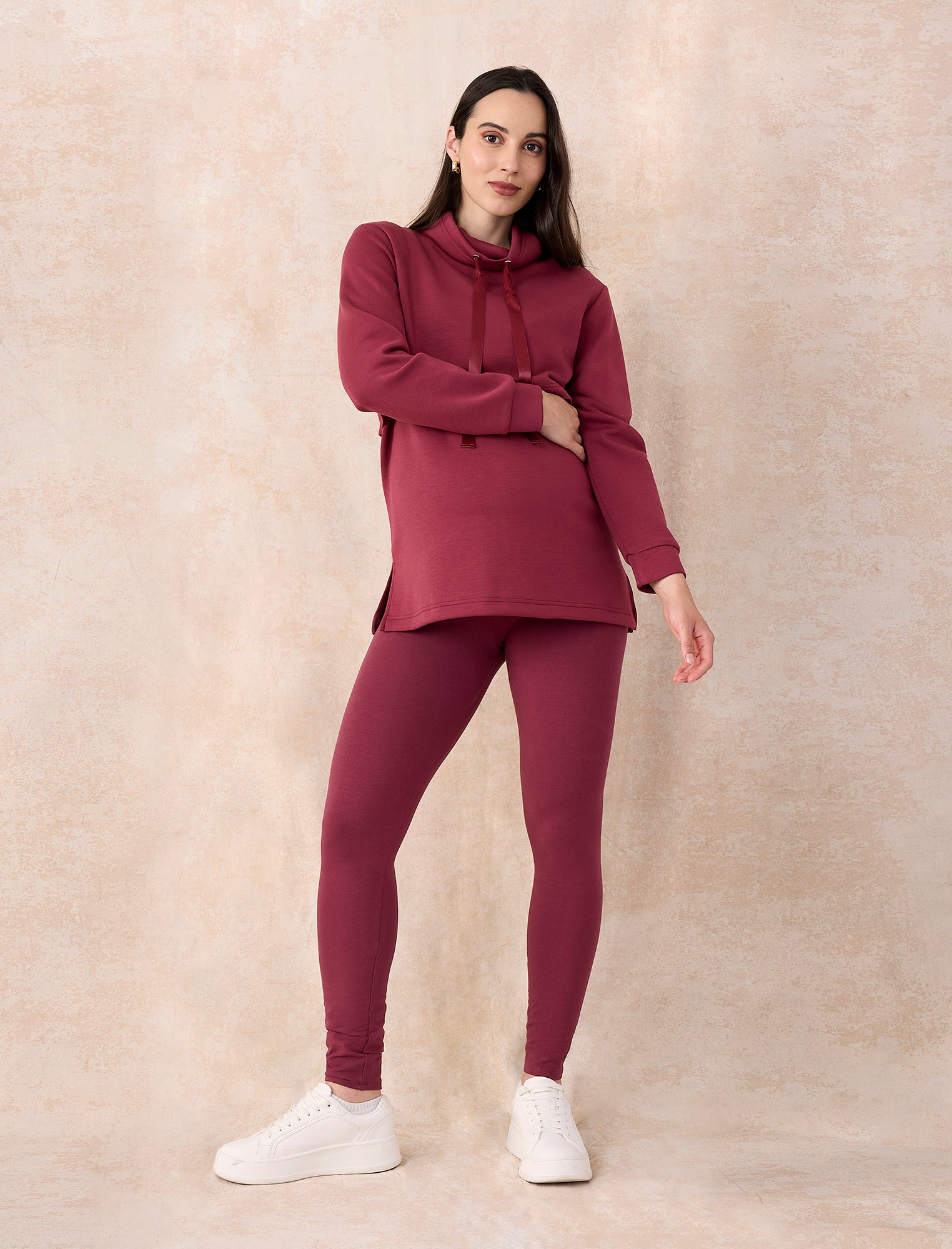 Pregnant woman wearing a maroon tracksuit with white shoes on a beige background