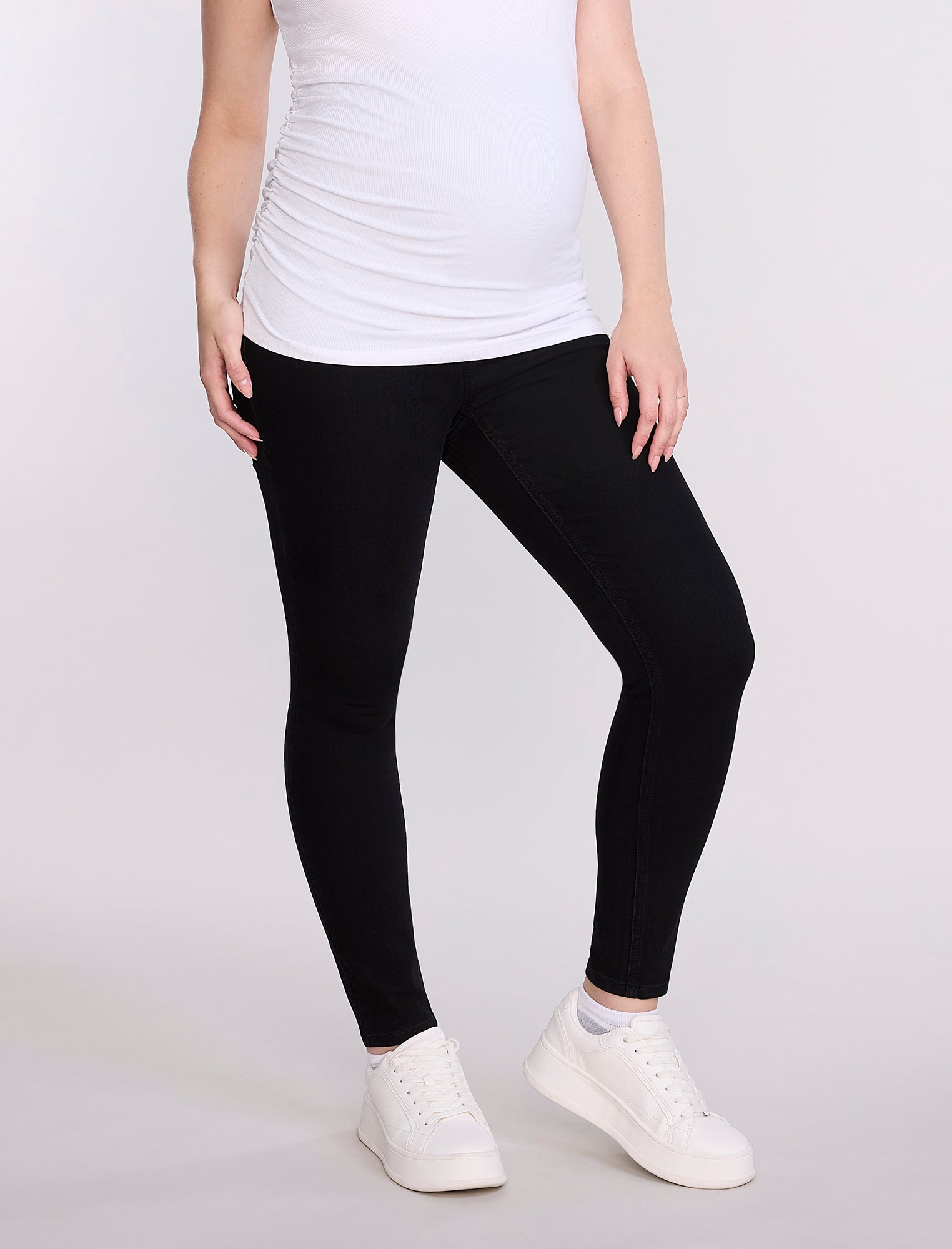 Pregnant woman wearing black leggings and a white top on a plain background
