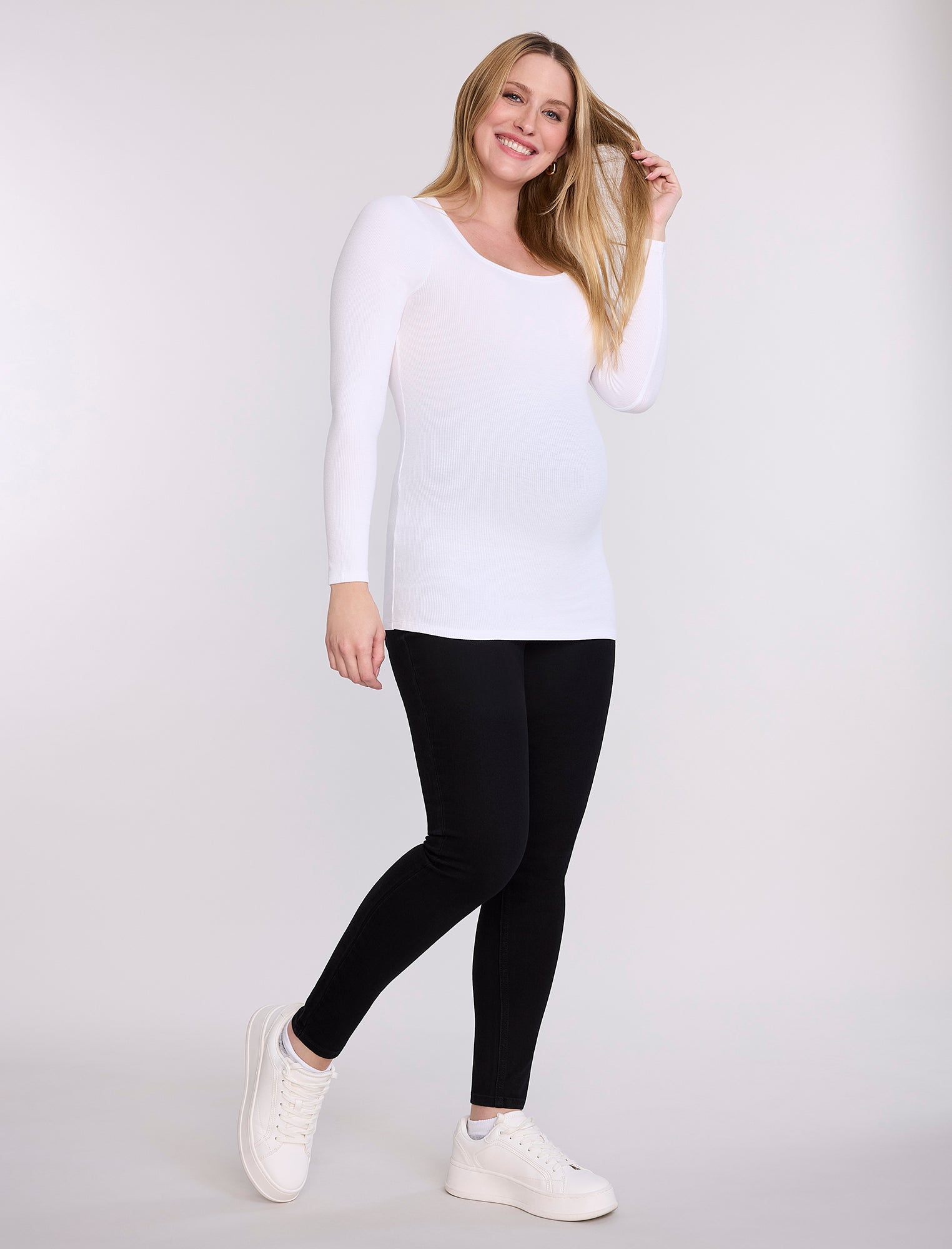 Pregnant woman wearing black leggings and a white top on a plain background