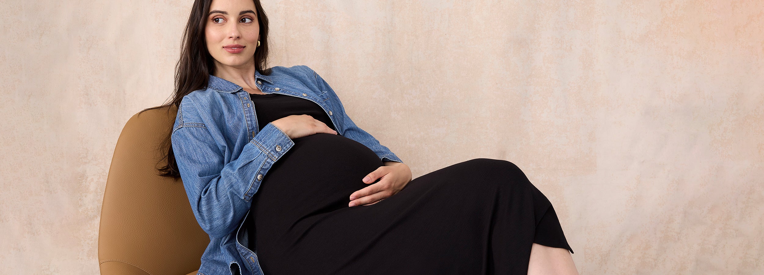 Pregnant woman sitting on a couch wearing a blue denim shirt over a black dress.