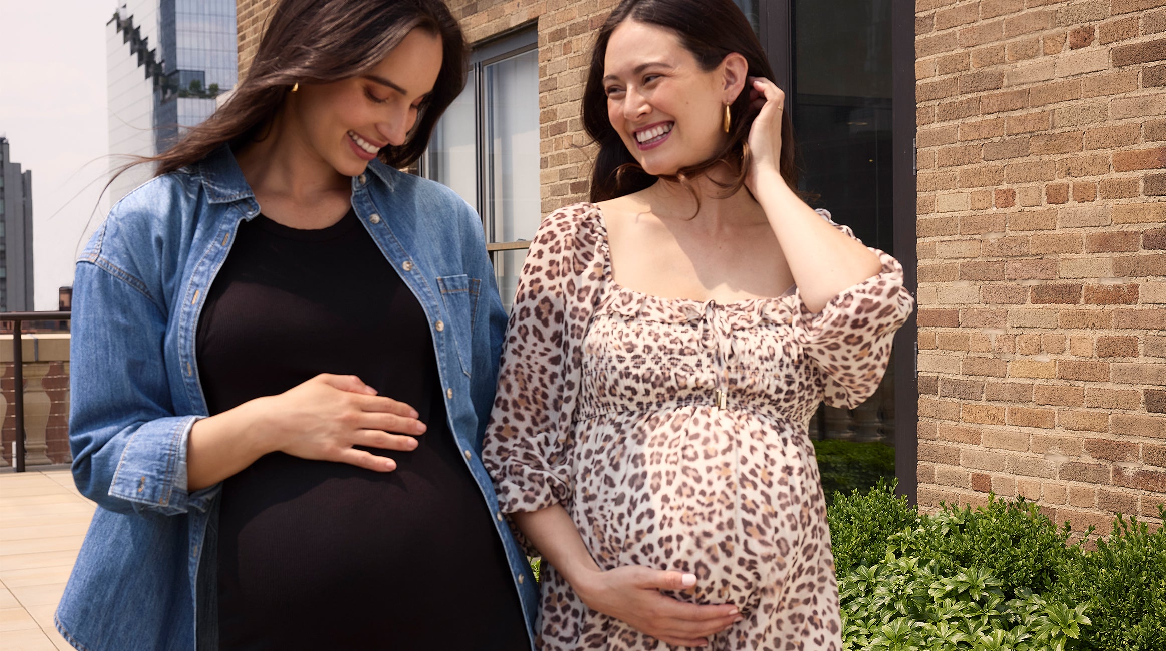 Two pregnant women standing outdoors, smiling and touching their bellies.
